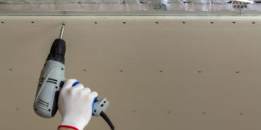 Close-up of constructor worker hand in protective glove with electrical cordless screwdriver connecting suspended drywall ceiling to metal frame. Renovation, construction and DIY concept.